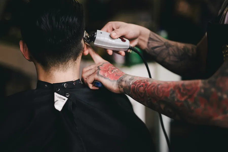 A tattooed barber running clippers along the neckline of a client in a black cape