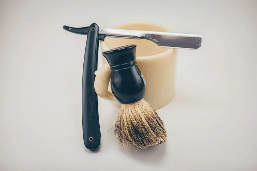 A straight razor and a shaving brush leaning on a cream shaving mug
