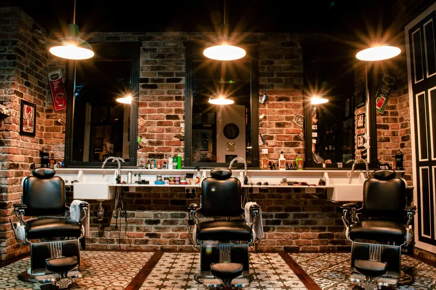 Three black leather barber chairs in front of mirrors on an exposed brick wall under pendant lamps